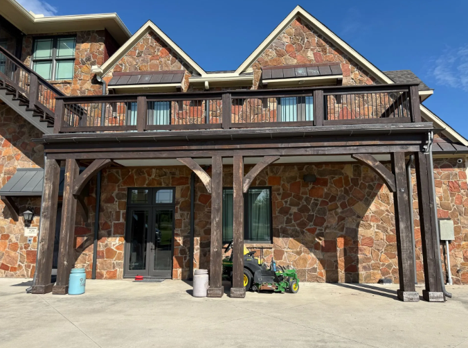 Brown wooden pergola-like structure attached to a stone-clad building; blue sky overhead.