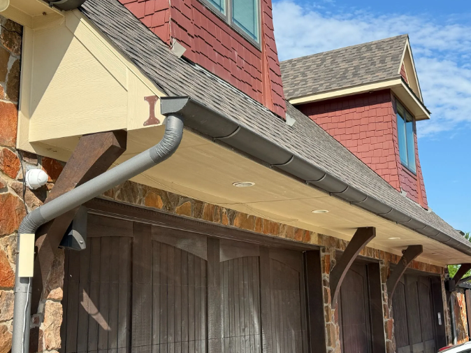 Gray gutters on a building with stone and wood exterior, brown garage doors, and a red roof against a blue sky.