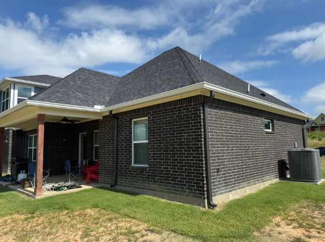 Dark brick house with a black roof, white gutters, and a green yard under a blue sky.