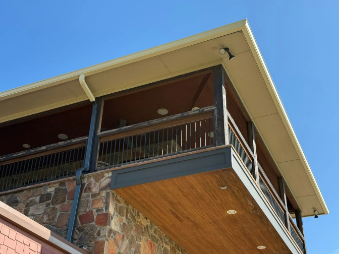 Wooden deck with railing, brown ceiling, stone facade, and beige gutter, against a blue sky.