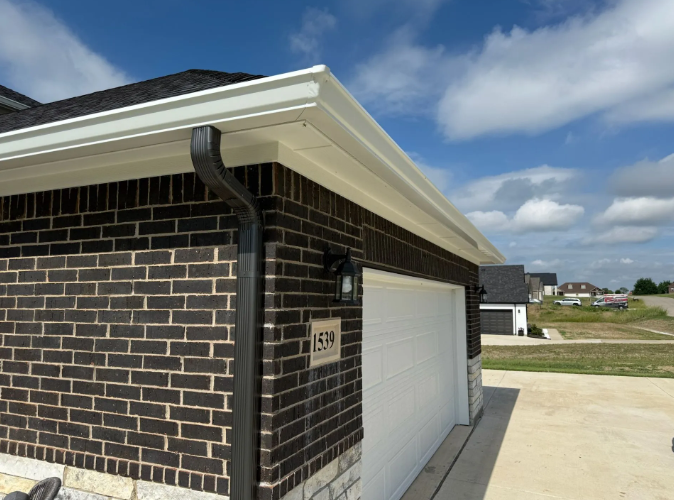 White gutters on a brick garage, black downspout. Sunny sky in background.