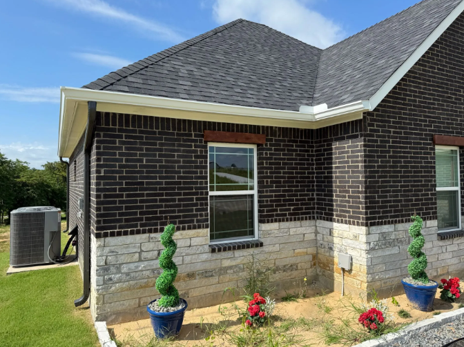 Brick house with dark roof and white trim. Green shrubs in blue pots, red flowers, and an AC unit.