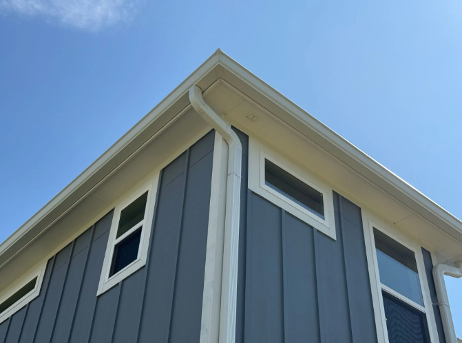 Exterior corner of a house with gray siding, white trim, gutters, and windows, against a blue sky.