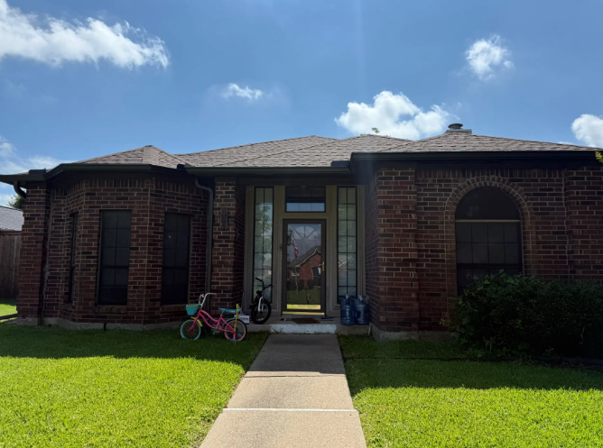 Brick house with a walkway and green lawn, blue sky and clouds. A small pink bike on the lawn.