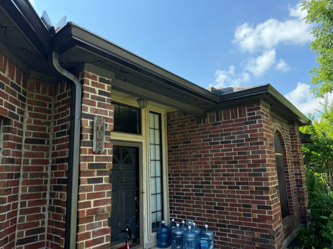 Brick house with dark gutters, doorway, and blue sky. Water jugs are visible near the door.