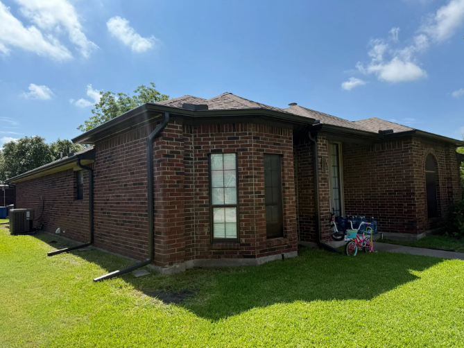 Brick house with brown roof, green lawn, blue sky, and black gutters.