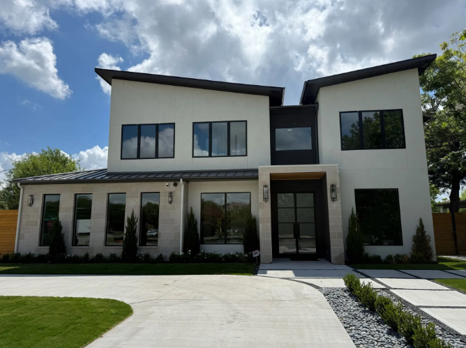 Modern two-story house with light-colored stucco, dark windows, and a black front door. Blue sky.
