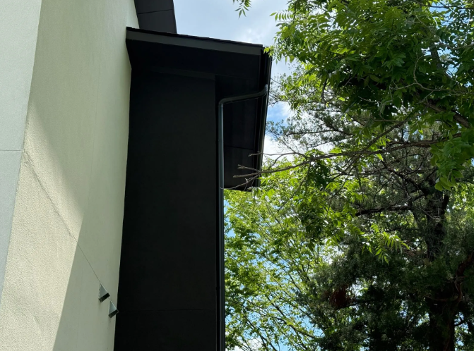 Black rectangular exterior architectural feature attached to a light-colored wall with visible gutter and tree backdrop.
