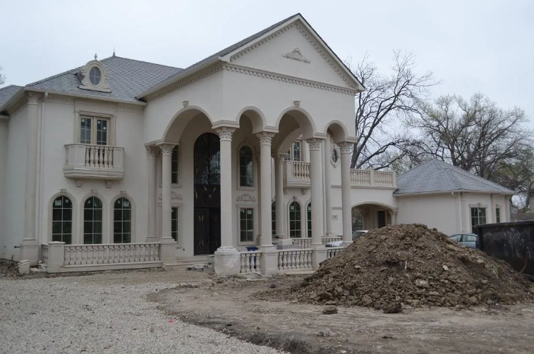 Two-story light-colored house with columns and a small detached building, with a dirt pile in front.