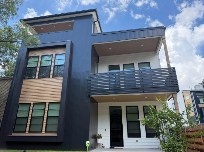 Modern three-story house with dark blue and white exterior, balcony, and contrasting wood detail. Blue sky with clouds.