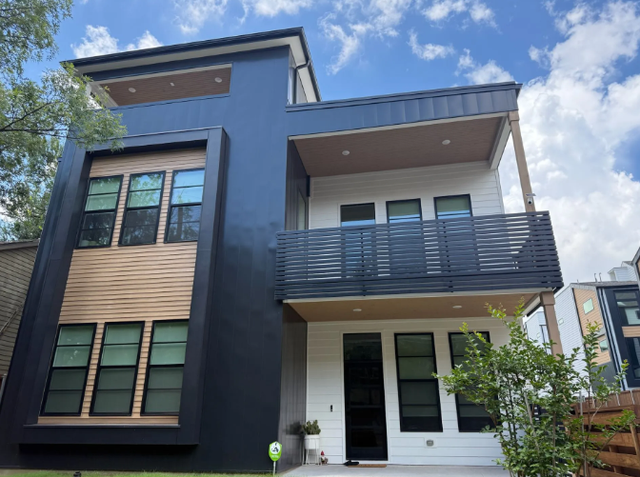 Modern three-story house with dark blue and white exterior, balcony, and contrasting wood detail. Blue sky with clouds.