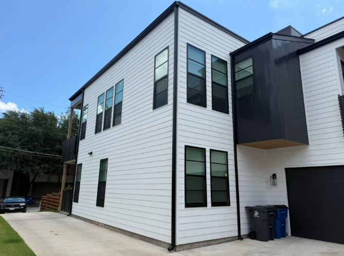 Modern two-story house with white siding, black trim, and large windows. Driveway and trash bins in view.