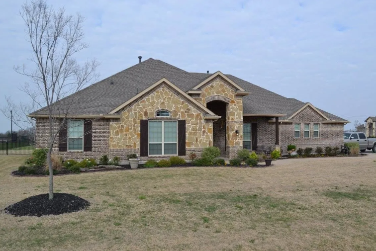 Tan brick and stone house with brown roof and landscaping under a cloudy sky.