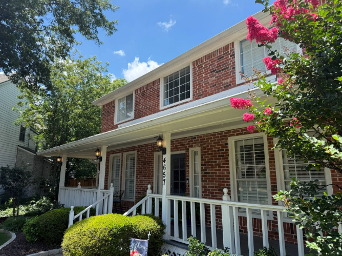 Two-story brick house with a porch. White trim, blue sky, and pink flowers.