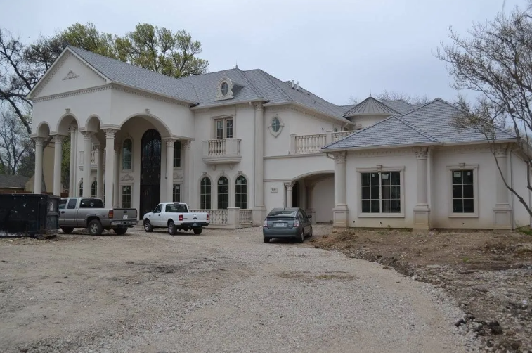 Large white mansion under construction with multiple columns, trucks parked in front, gravel driveway.