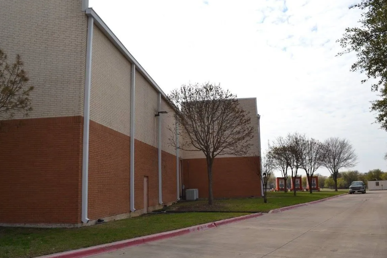 Brick building with brown and tan sections, next to a sidewalk and grassy area with trees.