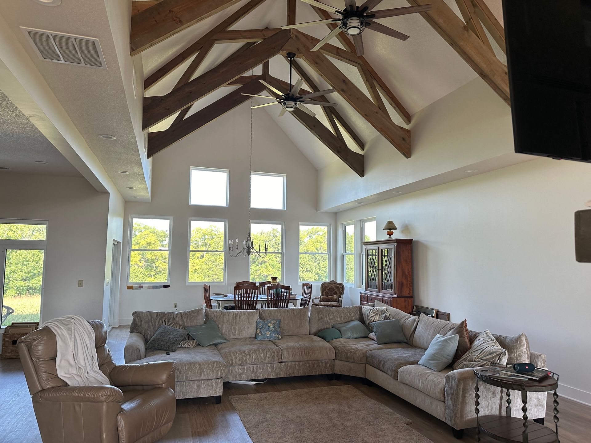 Spacious living room with high ceiling, exposed wooden beams, and large windows. A sectional sofa and armchair are in the foreground.