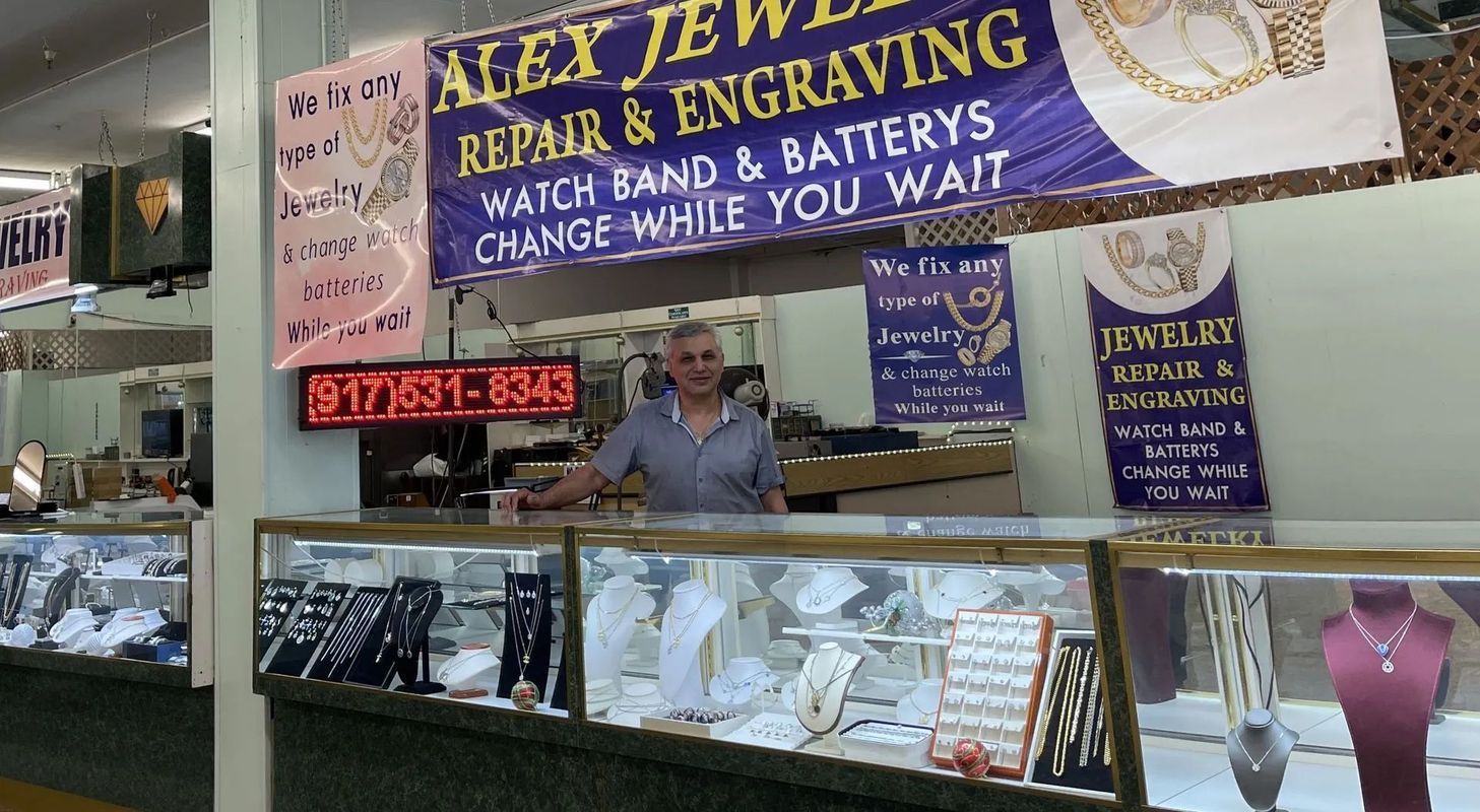 Jewelry store with a person behind the counter. Sign: Alex Jewelry, repair, engraving. Cases display jewelry.
