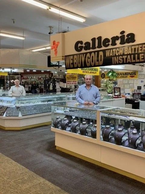 Jewelry store interior with two employees behind glass display cases filled with jewelry. Sign reads 
