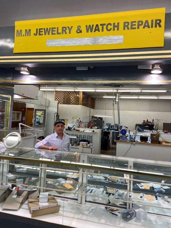 Man stands at a jewelry repair shop counter, under a yellow sign: M.M. JEWELRY & WATCH REPAIR. Shop interior visible.