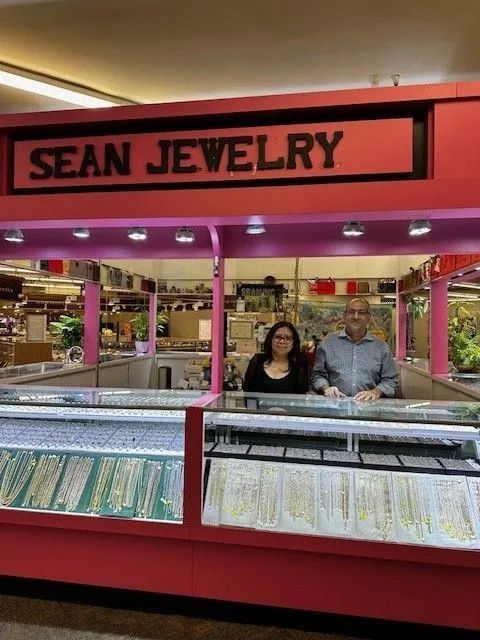 People at Sean Jewelry counter, inside a shop. Pink and red sign. Display cases with jewelry.