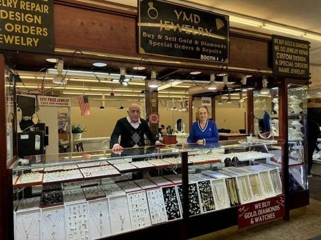 Two people stand behind a jewelry store counter filled with displays. Signage reads 