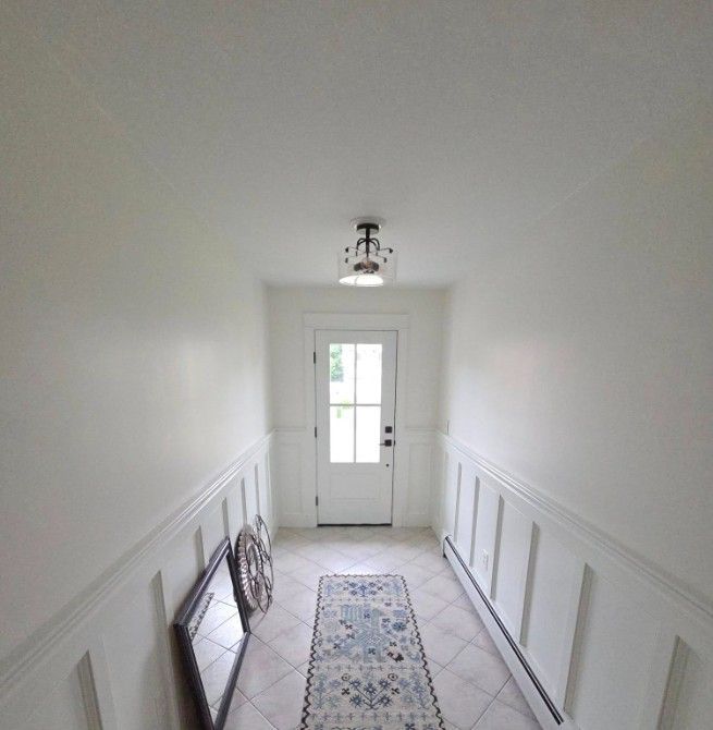 White hallway with wainscoting, door at the end, rug, and a light fixture.
