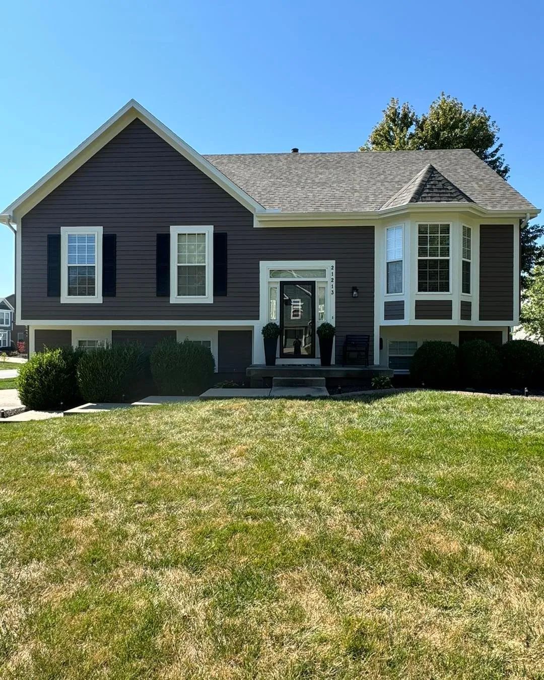 Brown house with white trim, black shutters, and green bushes in front.