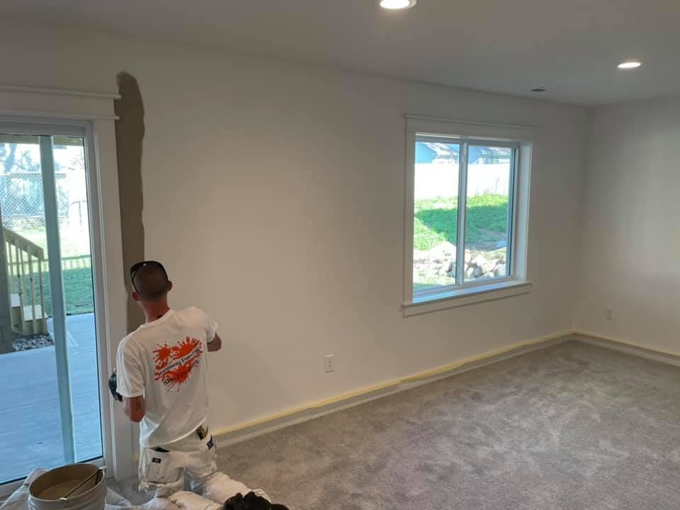 A painter in a white shirt and pants taping a wall, prepping to paint a room with a sliding door and a window.