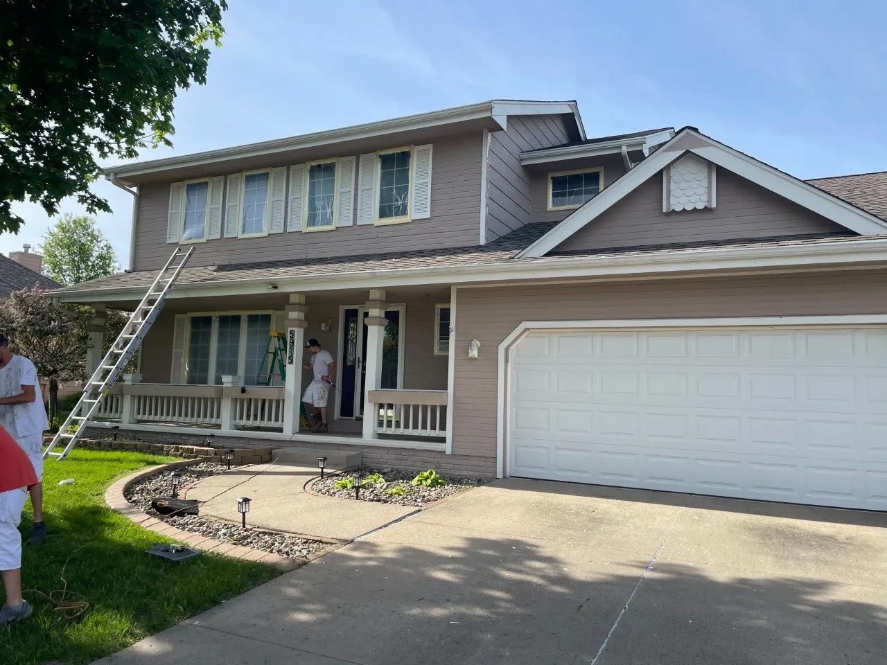 Two-story house being painted; tan siding, white trim, ladder, workers, sunny day.