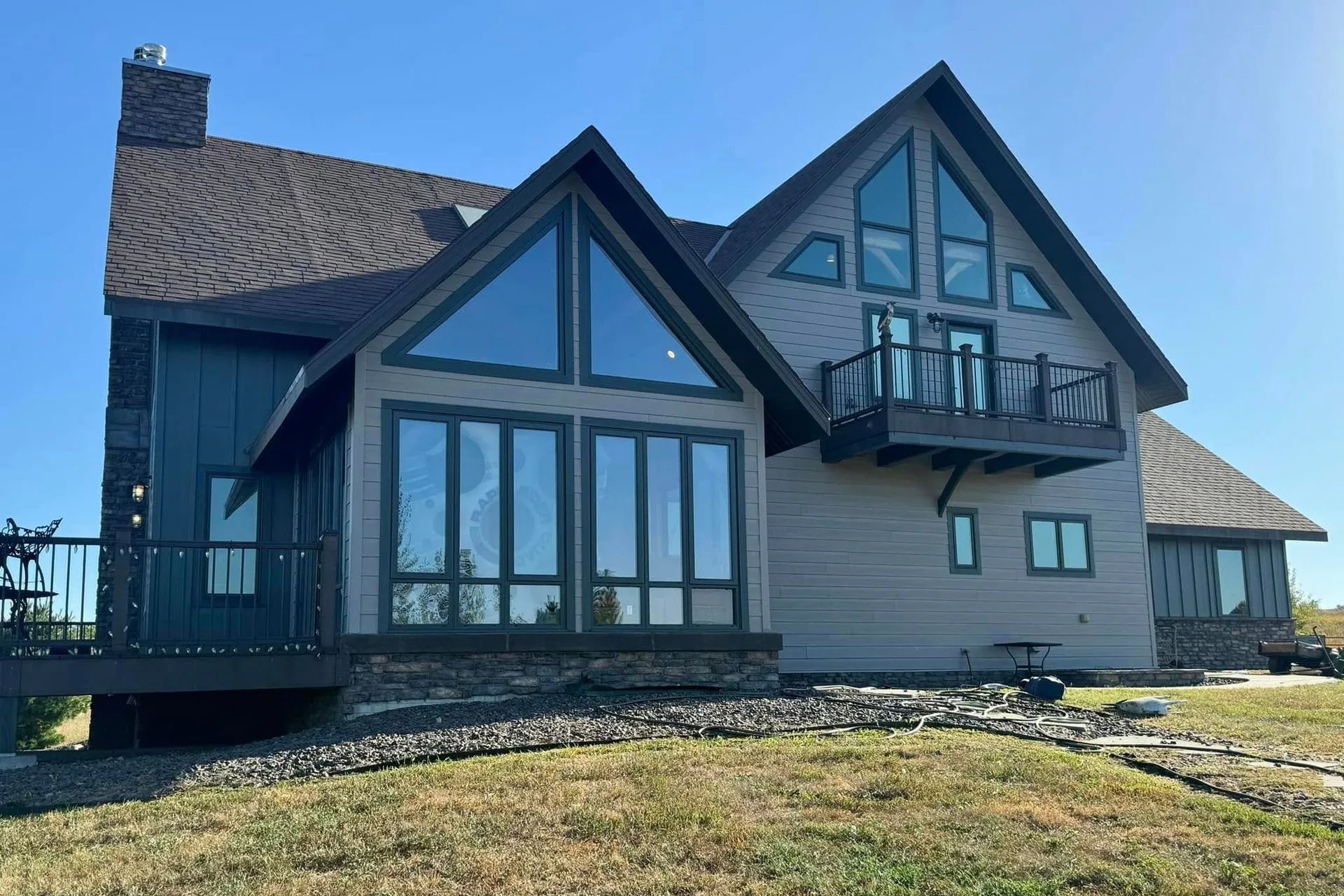 Two-story house with large windows and a deck, brown roof and siding, set against a bright blue sky.