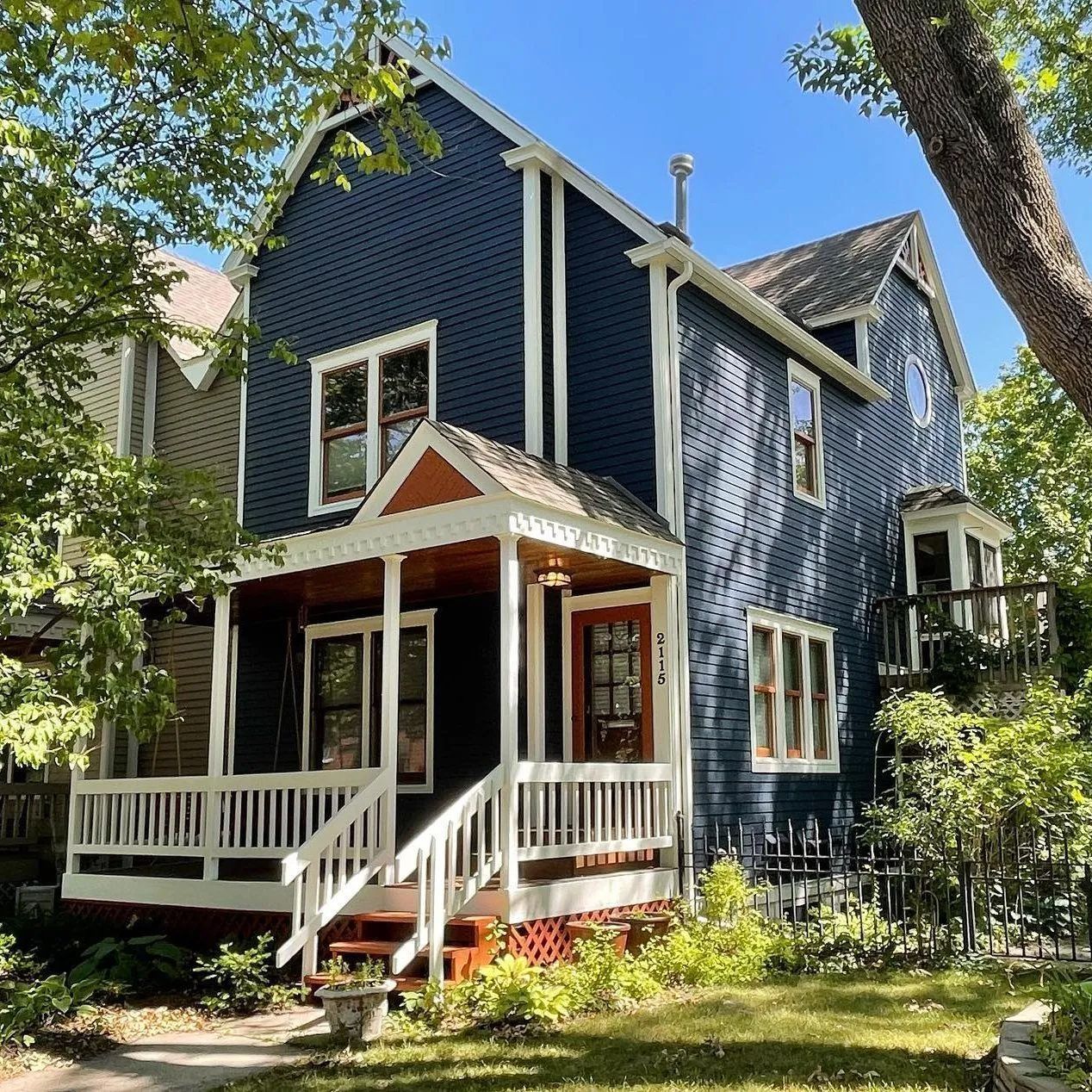 Blue two-story house with a porch, white trim, and a brown door. Lush green trees surround it.