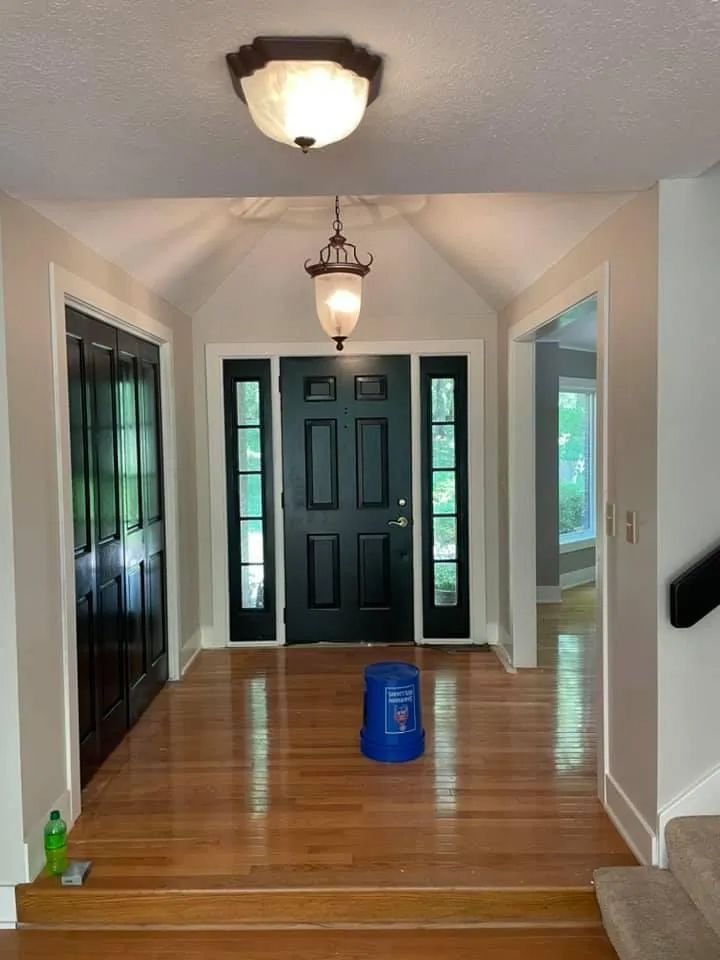 Entryway with wooden floors, black door, and light fixtures. Blue bucket on floor.