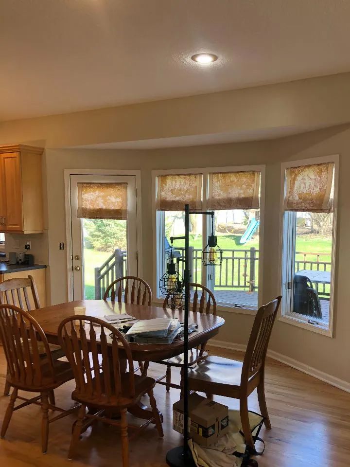 Dining room with wooden table and chairs, curved windows with shades overlooking a backyard.