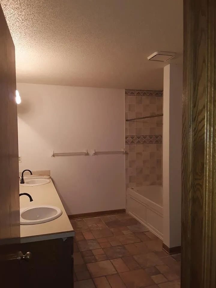 Bathroom with double sinks, tub with tile surround, brown floor, and towel racks.