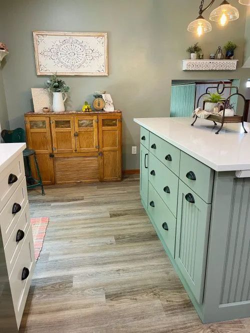 A farmhouse kitchen with a sage green island and cabinets, wooden floors, and antique furniture.