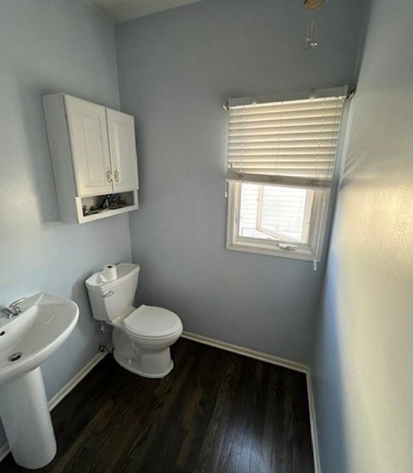 Small bathroom with blue walls, white toilet, sink, cabinet, and dark wood-look flooring. Window with blinds.