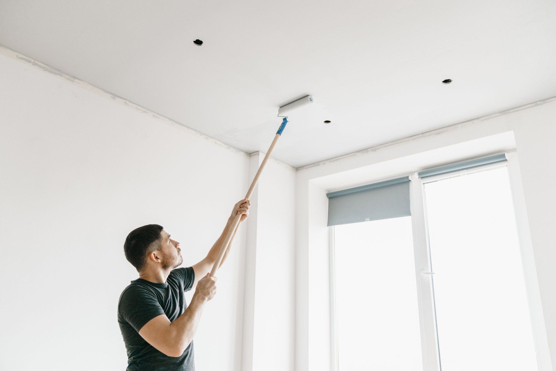 Man paints a white ceiling with a roller, standing in a room with a window.