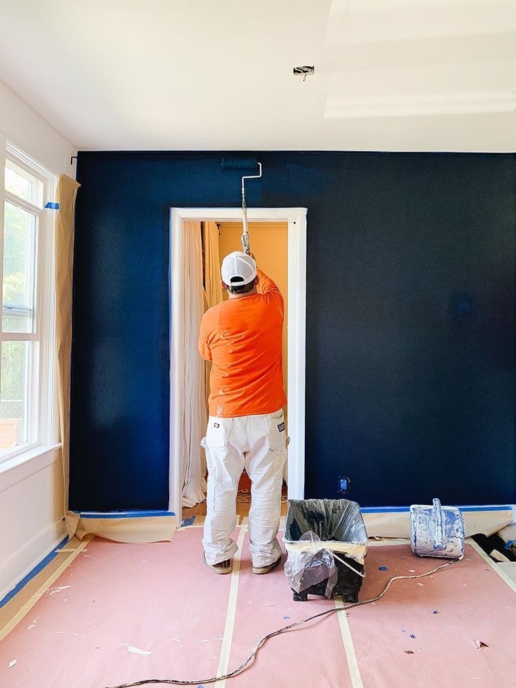 Man painting a dark blue accent wall in a room, wearing orange shirt and white pants.