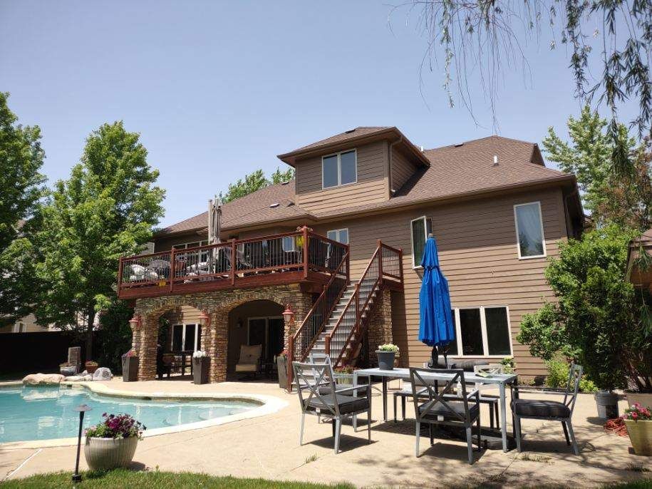 Two-story house with deck, pool, and patio furniture. Brown siding, blue umbrella, sunny day.