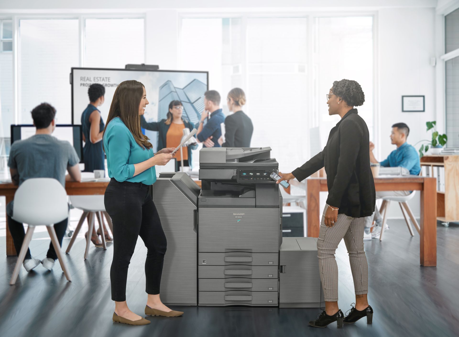 Two women are standing next to a printer in an office.