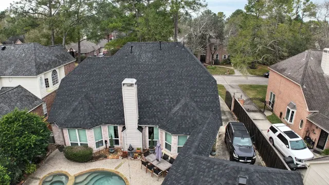 Aerial view of a house with a dark roof, white chimney, and parked cars in a suburban neighborhood.