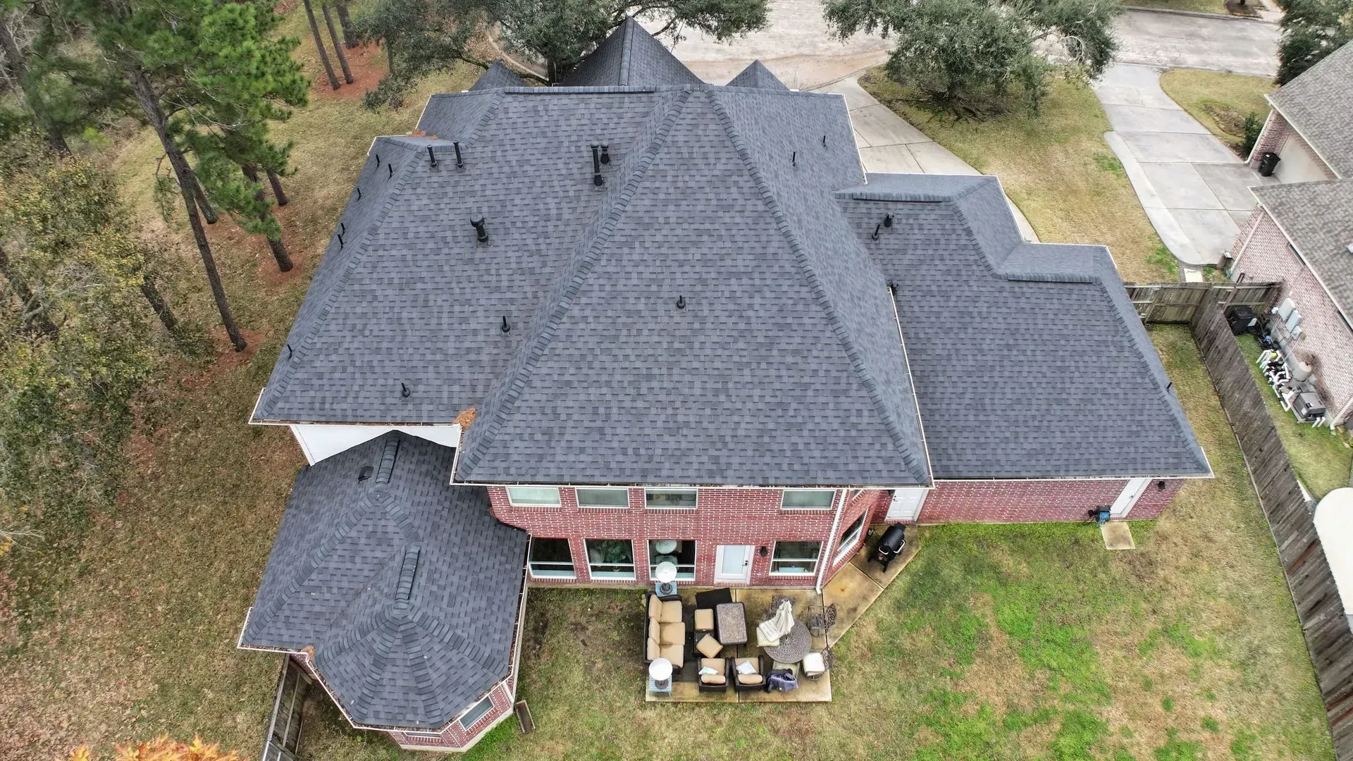 Aerial view of a large house with a dark gray roof, brick exterior, and a patio in a wooded yard.