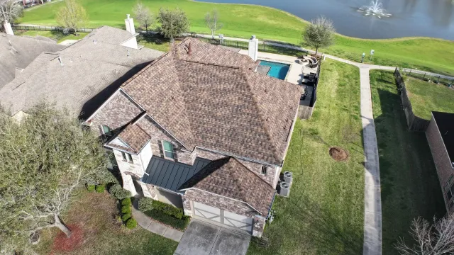 Aerial view of a tiled-roof house with a driveway, lawn, and trees beside a pond.