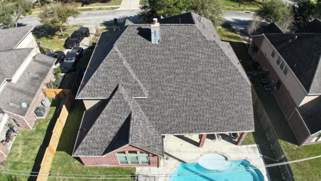 Aerial view of a suburban house with a dark shingle roof and backyard swimming pool.