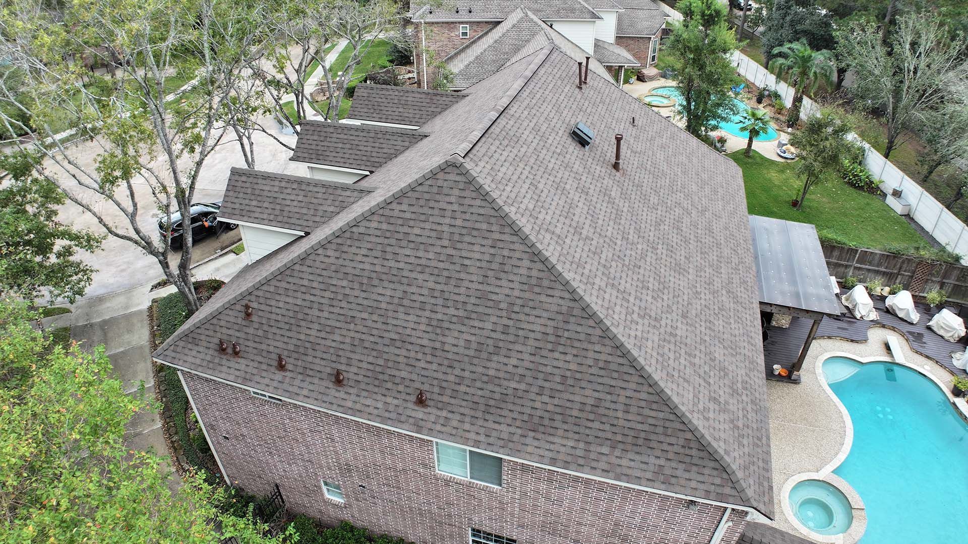 Aerial view of a house with a dark roof and backyard pool surrounded by trees and a fence