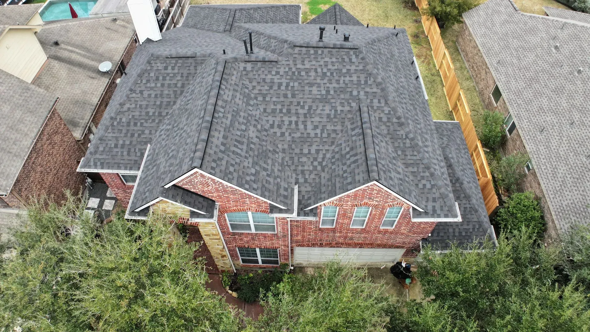 Aerial view of a brick house with a gray shingled roof and surrounding trees and neighboring homes