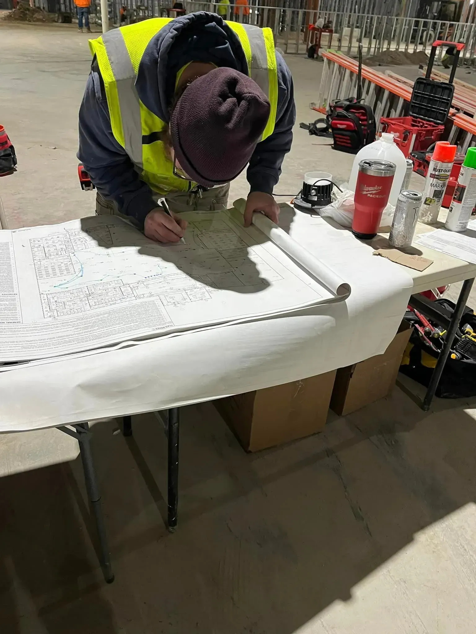 A person in a high-visibility vest and knit cap studies large architectural blueprints on a folding table at a job site.