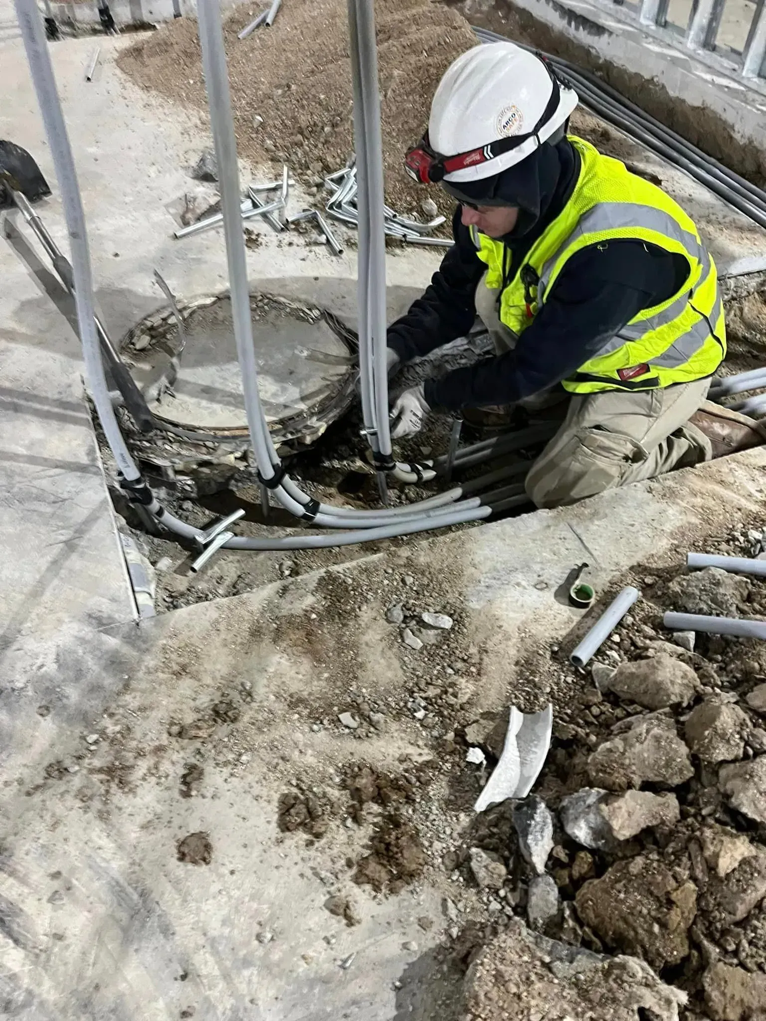 A worker in a hard hat and high-visibility vest inspects exposed electrical conduits at an outdoor construction site.