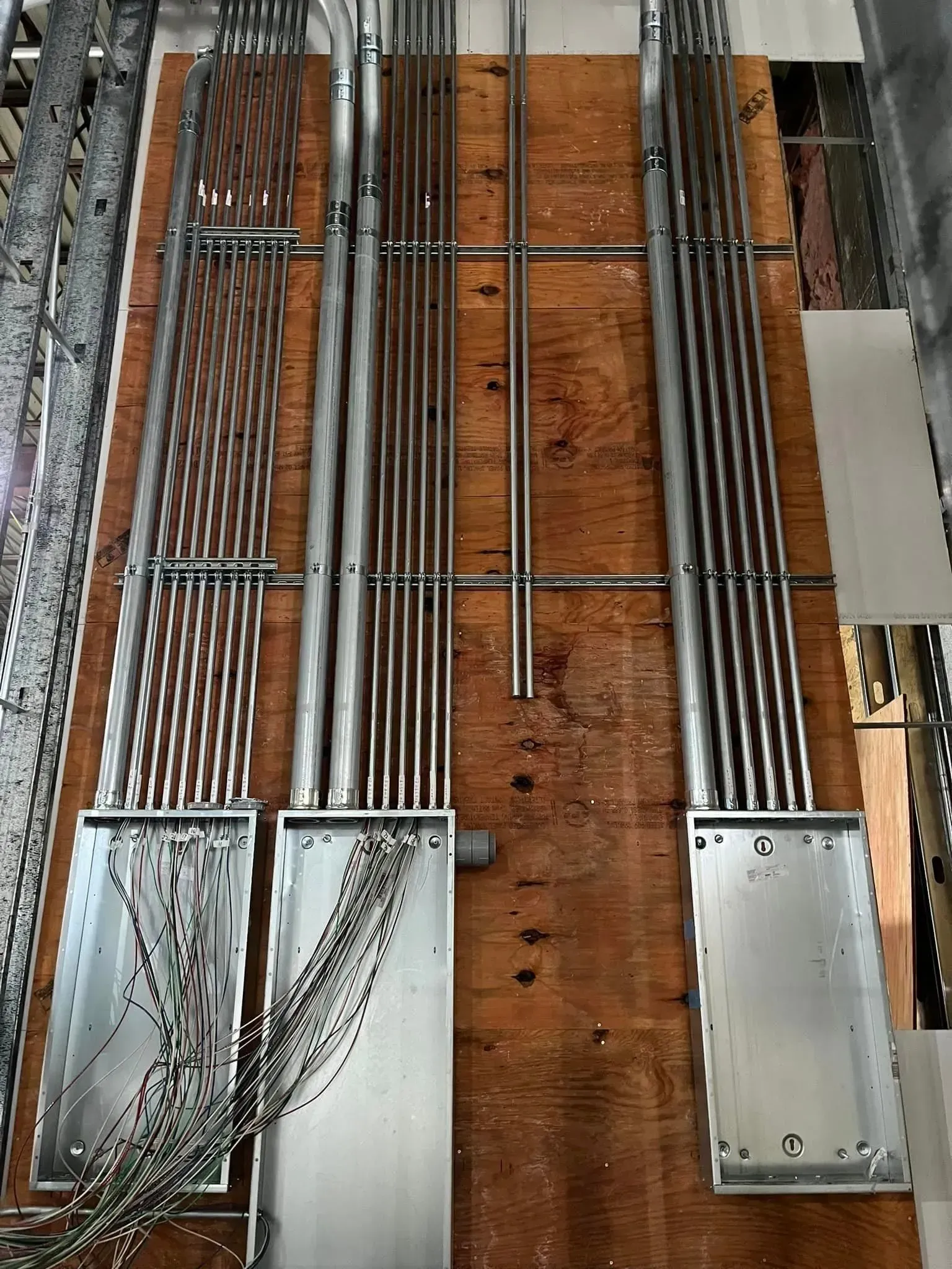 Three metal electrical boxes mounted on plywood with multiple conduit pipes running into them, some with exposed wires.